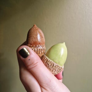 May include: Two small acorn-shaped ceramic ornaments. One acorn features a glossy brown nut with a textured, carved tan cap. The second acorn has a smooth, glossy light green nut paired with a matching textured, carved tan cap.