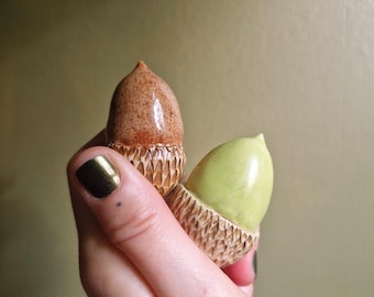 May include: Two small acorn-shaped ceramic ornaments. One acorn features a glossy brown nut with a textured, carved tan cap. The second acorn has a smooth, glossy light green nut paired with a matching textured, carved tan cap.