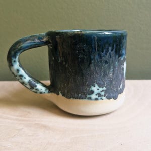 May include: A handmade ceramic mug with a unique glaze. The mug features a dark blue and black exterior with a cream-coloured base. The handle is a lighter blue with speckled detailing. The mug is approximately 10 cm tall.