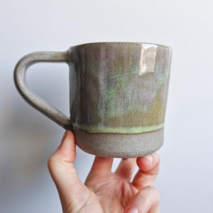 May include: A ceramic mug with a light grey glaze and a green and purple iridescent finish. The mug has a rounded handle and a slightly flared rim.
