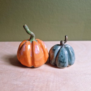 May include: Two ceramic pumpkin figurines with glossy glazes. The larger pumpkin is bright orange with a green stem, while the smaller pumpkin is a mottled blue-green colour with a dark brown stem. Both have distinct vertical ridges and curved stems.