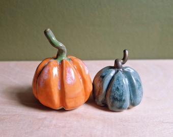 May include: Two ceramic pumpkin figurines with glossy glazes. The larger pumpkin is bright orange with a green stem, while the smaller pumpkin is a mottled blue-green colour with a dark brown stem. Both have distinct vertical ridges and curved stems.