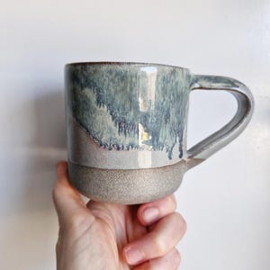 May include: A handcrafted ceramic mug with a distinctive glaze. The mug has a speckled, light grey base, a light grey mid-section, and a top section with a blue and grey glaze. The handle is light grey. The mug is held in a hand.