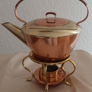Puede incluir: Una tetera de cobre y latón con un asa tejida se asienta sobre un soporte de cobre con un quemador. La tetera tiene una tapa con un borde de latón y una parte superior de cobre.