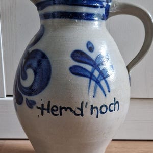 Könnte beinhalten: Steinzeugkrug mit Henkel, verziert mit blauen Blumenmustern und dem Text "Hemd'noch". Der Krug hat blaue Streifen um den Rand und ist hellbeige. Es ist eine traditionelle Art von Keramik.