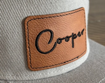 Peut inclure: Une casquette de baseball grise avec un patch en cuir marron qui dit "Cooper" en lettres cursives noires.