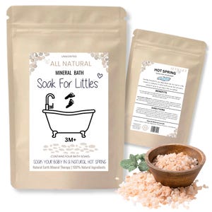 May include: Two beige pouches of mineral bath products for babies. The larger pouch features a label with a bathtub illustration and the text "Soak For Littles." The smaller pouch has "Hot Spring" text. A wooden bowl holds pink bath salts.