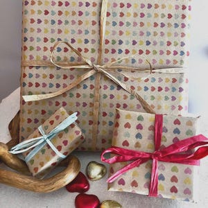 Puede incluir: Tres regalos envueltos con patrones de corazones coloridos sobre papel marr&oacute;n. El regalo m&aacute;s grande est&aacute; atado con un lazo de rafia. Un regalo m&aacute;s peque&ntilde;o est&aacute; atado con una cinta azul claro y otro con una cinta rosa. Chocolates en forma de coraz&oacute;n est&aacute;n esparcidos alrededor de los regalos.