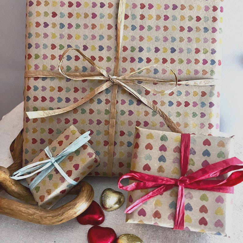Puede incluir: Tres regalos envueltos con patrones de corazones coloridos sobre papel marr&oacute;n. El regalo m&aacute;s grande est&aacute; atado con un lazo de rafia. Un regalo m&aacute;s peque&ntilde;o est&aacute; atado con una cinta azul claro y otro con una cinta rosa. Chocolates en forma de coraz&oacute;n est&aacute;n esparcidos alrededor de los regalos.