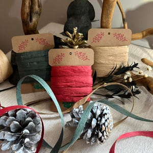 May include: A selection of craft supplies featuring spools of green, red, and natural raffia ribbon. Each spool has a kraft paper tag with a red floral design. The image also includes pine cones and decorative ribbon, ideal for crafting and gift wrapping.