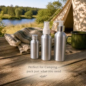 May include: Three silver aluminum bottles with different tops, including a spray bottle and a pump bottle, are arranged on a wooden surface. A folded towel and a green enamel mug are nearby. The text reads, "Perfect for Camping - pack just what you need."