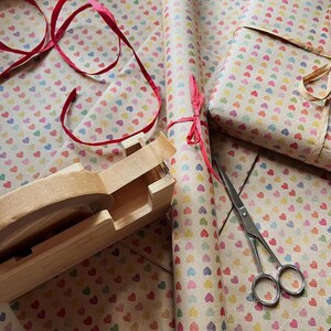 Puede incluir: Suministros para envolver regalos, incluyendo rollos de papel con peque&ntilde;os corazones coloridos, un regalo envuelto, tijeras y un dispensador de cinta adhesiva. Una cinta roja a&ntilde;ade un toque decorativo a la escena.