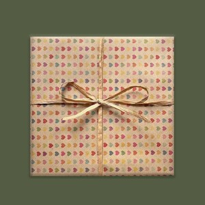 Puede incluir: Un paquete cuadrado envuelto en papel marr&oacute;n con un patr&oacute;n de corazones coloridos. Est&aacute; atado con una cinta y lazo de rafia natural. El fondo es de color verde oliva liso.