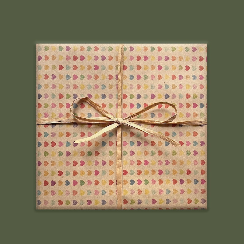 Puede incluir: Un paquete cuadrado envuelto en papel marr&oacute;n con un patr&oacute;n de corazones coloridos. Est&aacute; atado con una cinta y lazo de rafia natural. El fondo es de color verde oliva liso.