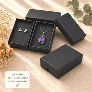 May include: Black jewellery boxes displaying a necklace and earrings. The necklace has a gold chain and a purple pendant. The earrings are silver with a Celtic knot design. The boxes are on a beige surface. Text on a label reads: UK MADE, RECYCLED CARD, READY ASSEMBLED.