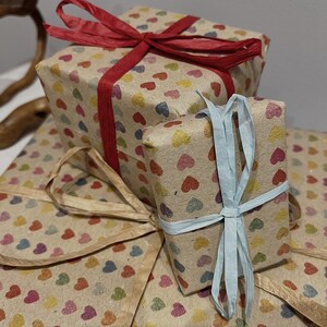 Puede incluir: Varias cajas de regalo envueltas en papel marr&oacute;n con patrones de corazones coloridos. Las cajas est&aacute;n atadas con cintas rojas y azul claro. Los regalos est&aacute;n apilados.