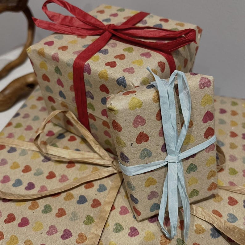 Puede incluir: Varias cajas de regalo envueltas en papel marr&oacute;n con patrones de corazones coloridos. Las cajas est&aacute;n atadas con cintas rojas y azul claro. Los regalos est&aacute;n apilados.