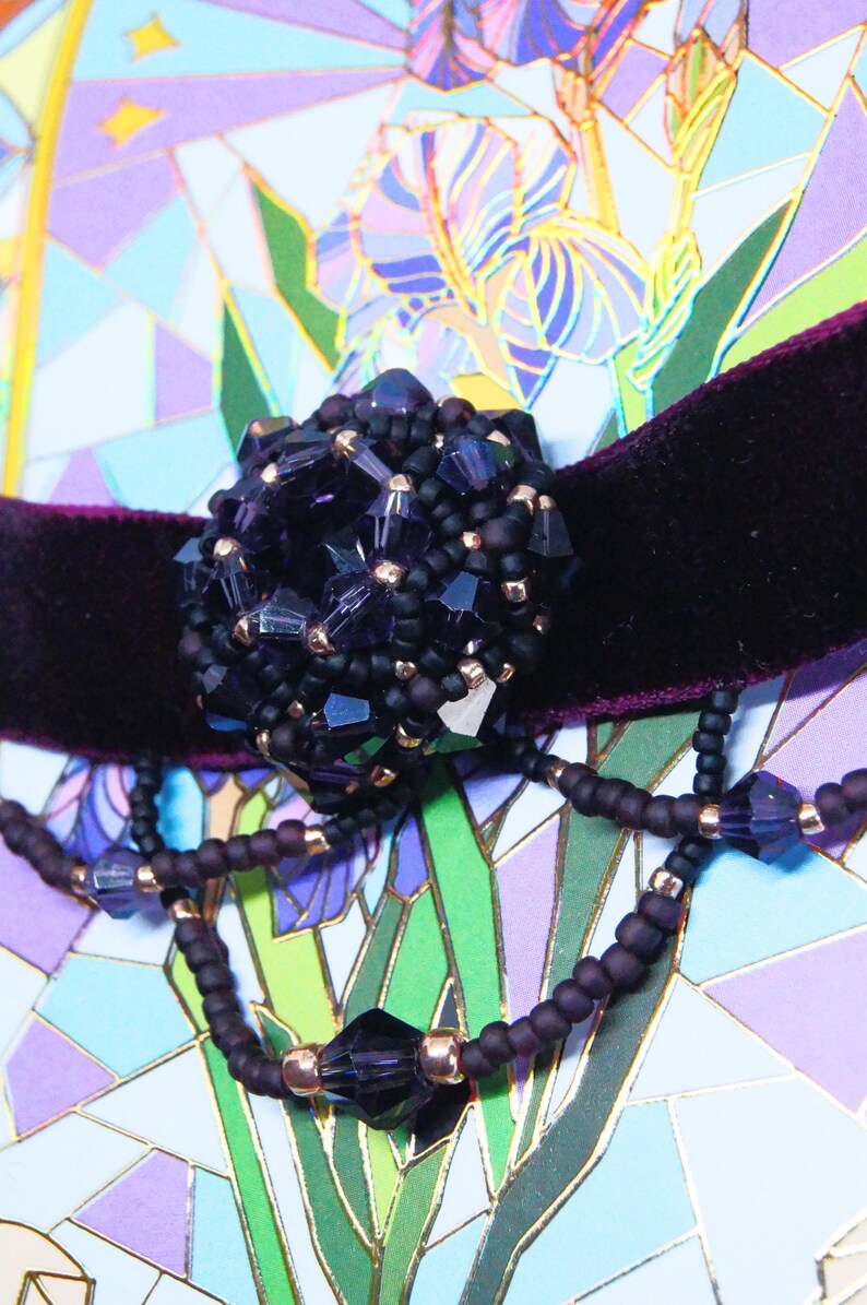 May include: A close-up of a dark purple beaded choker necklace with a large, clustered centerpiece. The centerpiece and necklace feature faceted beads and small gold-colored accents. The necklace sits on a dark purple velvet band.
