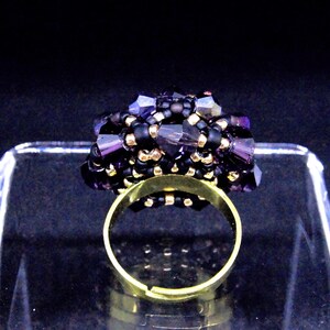 May include: A handmade ring featuring a cluster of dark purple and black beads. The ring has a gold-colored adjustable band. The beaded cluster is the focal point, with a mix of small black beads and larger, faceted purple beads.