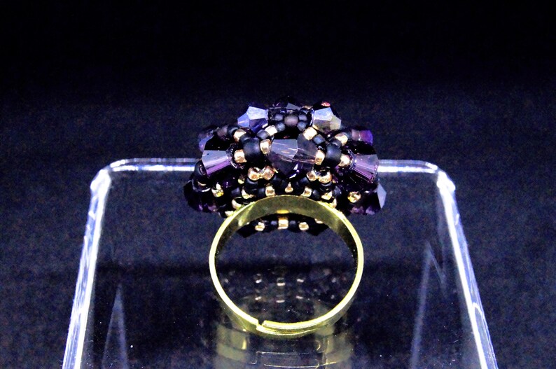May include: A handmade ring featuring a cluster of dark purple and black beads. The ring has a gold-colored adjustable band. The beaded cluster is the focal point, with a mix of small black beads and larger, faceted purple beads.