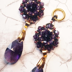 May include: A pair of handcrafted earrings featuring a cluster of deep purple beads and a teardrop-shaped amethyst gemstone. The earrings have gold-colored accents and a gold hoop for the ear. The design is elegant and suitable for special occasions.