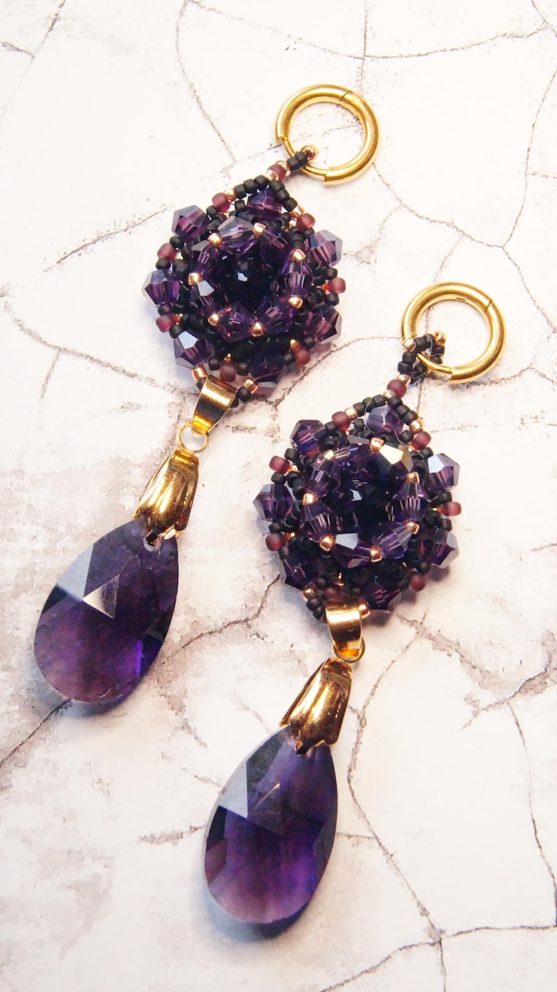 May include: A pair of handcrafted earrings featuring a cluster of deep purple beads and a teardrop-shaped amethyst gemstone. The earrings have gold-colored accents and a gold hoop for the ear. The design is elegant and suitable for special occasions.