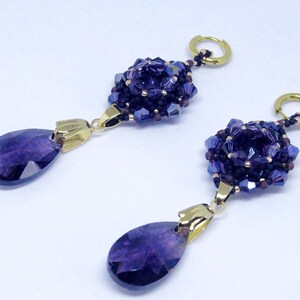 May include: A pair of dangle earrings featuring a cluster of dark purple beads and a teardrop-shaped purple gemstone. The earrings have gold-colored accents and a gold hoop closure. The jewelry is set against a white background.