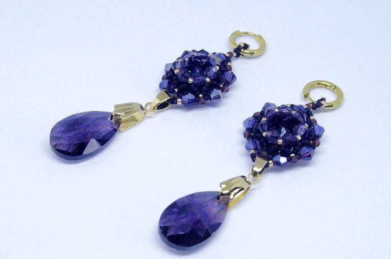 May include: A pair of dangle earrings featuring a cluster of dark purple beads and a teardrop-shaped purple gemstone. The earrings have gold-colored accents and a gold hoop closure. The jewelry is set against a white background.