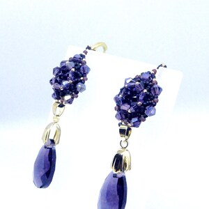 May include: A pair of dangle earrings featuring clusters of small, faceted purple beads. Each earring has a gold-colored cap and a large, teardrop-shaped purple gemstone. The earrings are displayed against a white background.