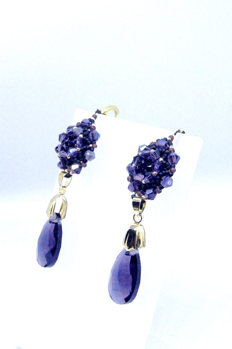 May include: A pair of dangle earrings featuring clusters of small, faceted purple beads. Each earring has a gold-colored cap and a large, teardrop-shaped purple gemstone. The earrings are displayed against a white background.