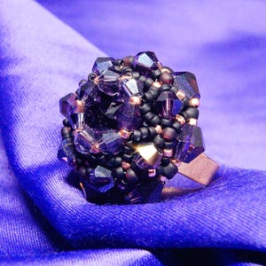 May include: A handcrafted ring featuring a cluster of faceted, dark purple crystals and black beads. The ring's design is set on a copper-colored band, creating a striking contrast against the deep purple hue. The ring is displayed on a blue fabric.