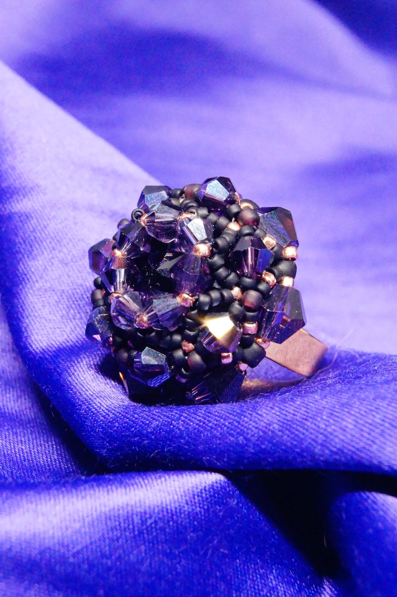 May include: A handcrafted ring featuring a cluster of faceted, dark purple crystals and black beads. The ring's design is set on a copper-colored band, creating a striking contrast against the deep purple hue. The ring is displayed on a blue fabric.