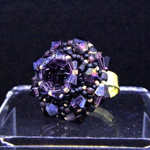 May include: A beaded ring featuring a cluster of purple and black beads. The ring has a gold-colored band and is displayed on a clear, angled surface against a dark background. The beads have a faceted, crystal-like appearance.