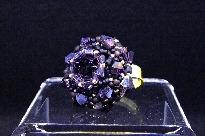 May include: A beaded ring featuring a cluster of purple and black beads. The ring has a gold-colored band and is displayed on a clear, angled surface against a dark background. The beads have a faceted, crystal-like appearance.