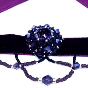 May include: A dark purple choker necklace with a black velvet band and a large, beaded centerpiece. The centerpiece is a cluster of dark blue and black beads. Dangling strands of beads add detail.
