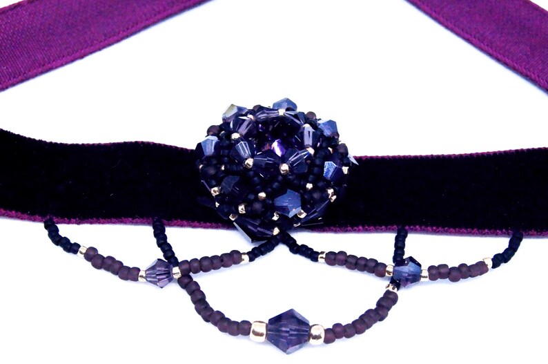 May include: A dark purple choker necklace with a black velvet band and a large, beaded centerpiece. The centerpiece is a cluster of dark blue and black beads. Dangling strands of beads add detail.