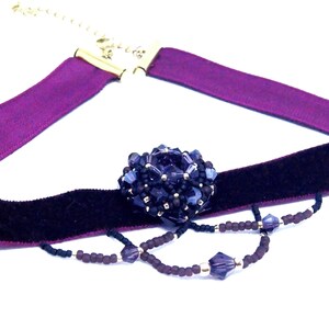 May include: A choker necklace featuring a dark purple velvet band and a lighter purple satin ribbon. The necklace has a central beaded embellishment in shades of blue and purple, with a delicate beaded chain.