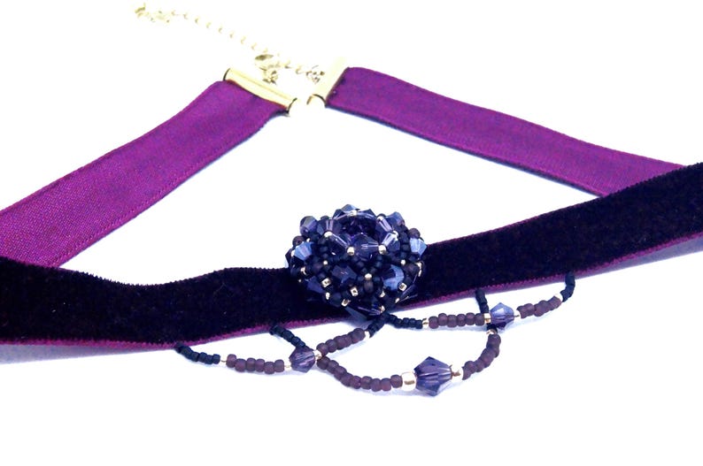 May include: A choker necklace featuring a dark purple velvet band and a lighter purple satin ribbon. The necklace has a central beaded embellishment in shades of blue and purple, with a delicate beaded chain.