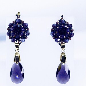 May include: A pair of elegant dangle earrings. Each earring features a cluster of dark purple beads and crystals, with a teardrop-shaped, faceted purple crystal pendant. Gold-colored metal accents connect the elements. The earrings are set against a white background.