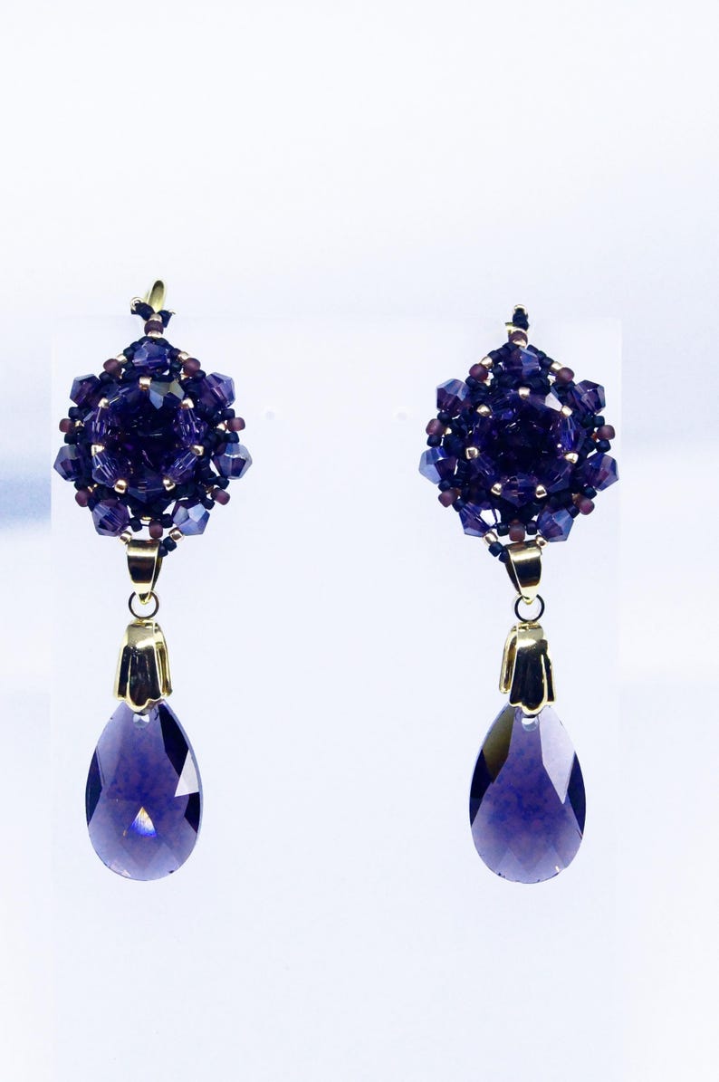 May include: A pair of elegant dangle earrings. Each earring features a cluster of dark purple beads and crystals, with a teardrop-shaped, faceted purple crystal pendant. Gold-colored metal accents connect the elements. The earrings are set against a white background.