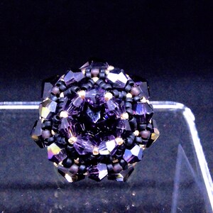 May include: A handcrafted ring featuring a central faceted purple crystal surrounded by smaller black and purple beads. The ring's design is geometric, with a dark background. The ring is a piece of jewelry.