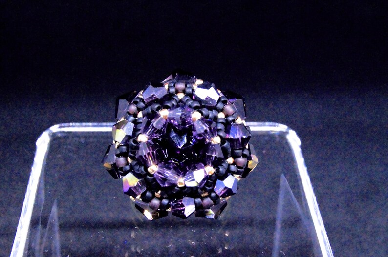 May include: A handcrafted ring featuring a central faceted purple crystal surrounded by smaller black and purple beads. The ring's design is geometric, with a dark background. The ring is a piece of jewelry.