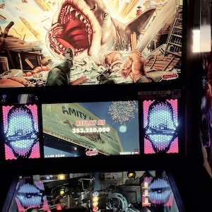 May include: A Jaws-themed pinball machine with a detailed playfield. The backboard features the movie's logo and a shark illustration. The machine has various lights and scoring displays, with the Amity Island sign at the top.