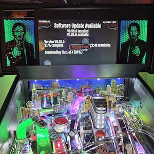 May include: A John Wick pinball machine with a cityscape playfield. The machine's display shows a software update in progress, with the text "Software Update Available". The game's logo is visible at the top.