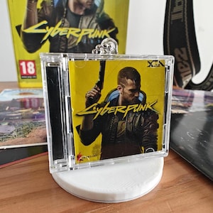 Può includere: Una custodia in plastica trasparente con una copertina di videogioco Cyberpunk 2077 gialla e nera. La copertina presenta un personaggio con una pistola e il testo "Cyberpunk".