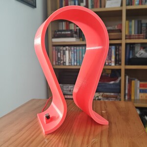 Op de afbeelding: Een felroze plastic hoofdtelefoonstandaard met een gebogen ontwerp. De standaard staat op een houten oppervlak.