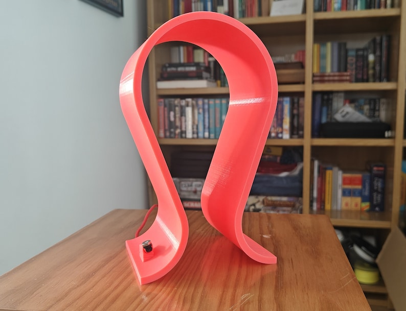 Op de afbeelding: Een felroze plastic hoofdtelefoonstandaard met een gebogen ontwerp. De standaard staat op een houten oppervlak.