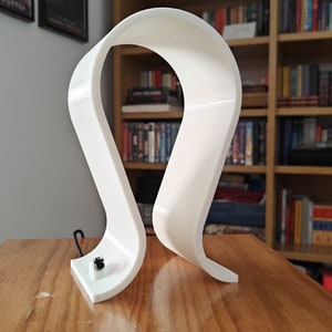 Op de afbeelding: Witte plastic hoofdtelefoonstandaard met een gebogen ontwerp. De standaard staat op een houten oppervlak.