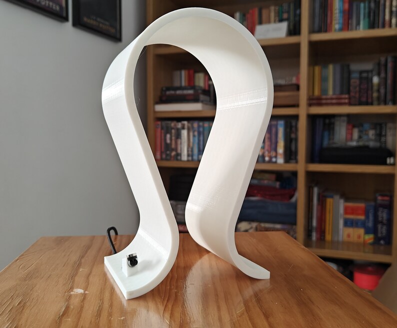 Op de afbeelding: Witte plastic hoofdtelefoonstandaard met een gebogen ontwerp. De standaard staat op een houten oppervlak.
