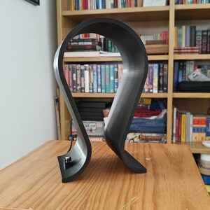 Op de afbeelding: Een zwarte plastic hoofdtelefoonstandaard met een gebogen ontwerp. De standaard staat op een houten oppervlak.